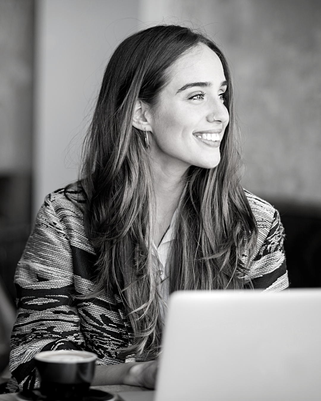 Black and white image of a young woman with long brown hair. She is smiling as she works from her laptop.