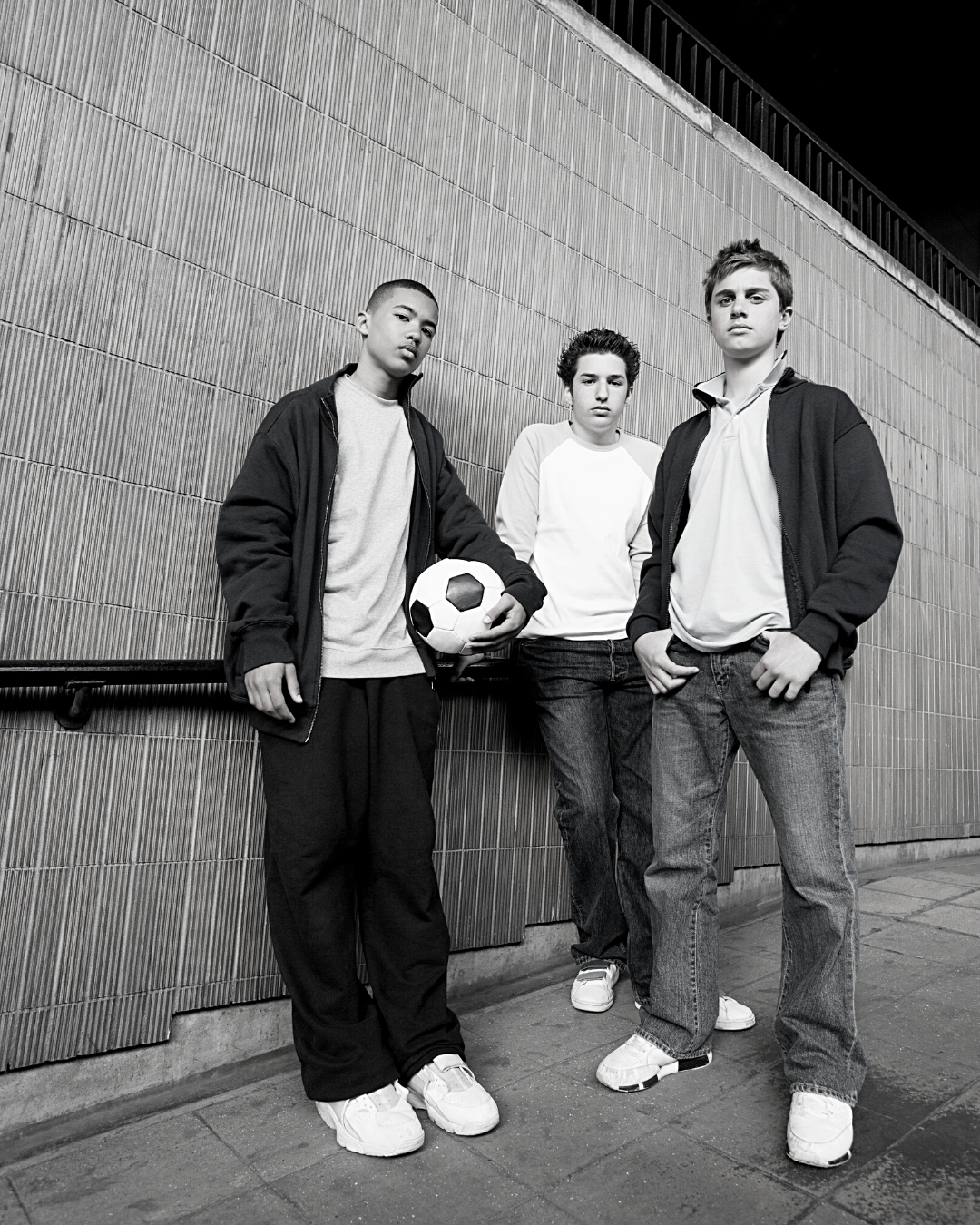 Three teen boys leaning against a wall, one is holding a soccer ball.