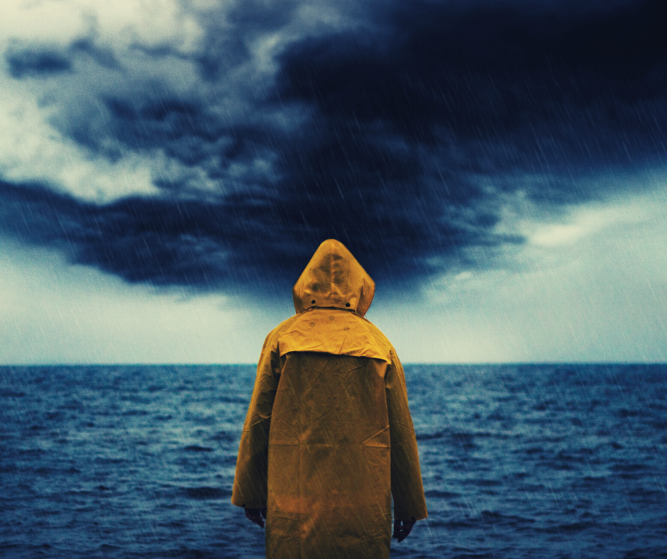 An individual with their back to the camera, wearing a yellow rain coat and staring out to see. Dark storm clouds are visible.